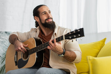 Obraz premium bearded hispanic musician looking away while playing guitar at home