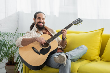happy latin man looking at camera while playing guitar on yellow sofa at home