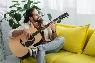 latin man playing guitar while sitting on yellow couch at home