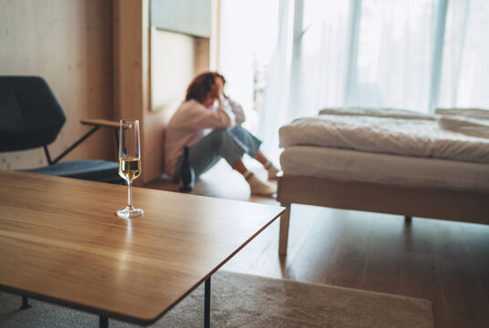 Wine glass on the table with the unfocused sad woman sitting on the floor in the bedroom with a bottle of alcohol. Mental health and alcoholism problems concept image.