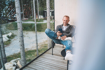 Fototapeta premium Smiling middle-aged man dressed open cardigan, jeans, and warm socks sitting on forest house balcony and using modern smartphone. Everyday lifestyle photo with modern devices concept image.