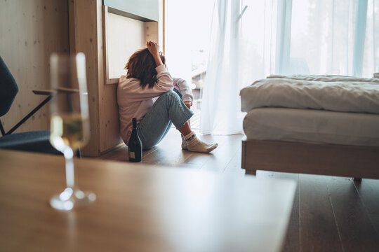Sad woman sitting on the floor in the bedroom with a bottle of alcohol. Unfocused wine glass on the foreground table. Mental health and alcoholism problems concept image.