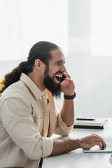 excited hispanic freelancer laughing while talking on cellphone near laptop