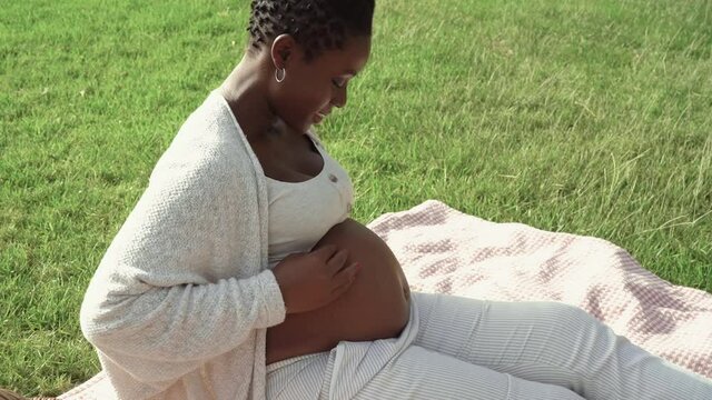 Young African woman caressing her pregnant belly in park - Maternity lifestyle concept