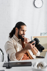 thoughtful hispanic man touching beard while looking at digital camera near blurred graphic tablet