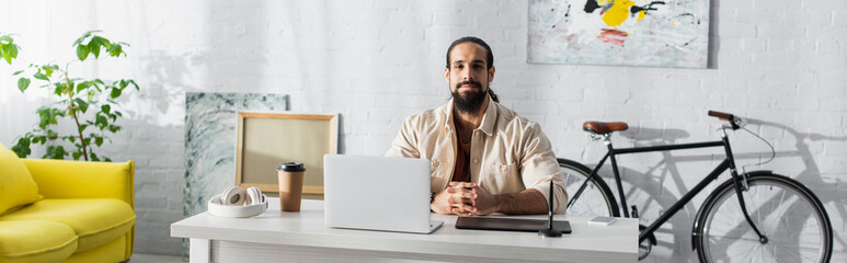 bearded latin freelancer sitting near laptop and graphic tablet in home studio, banner