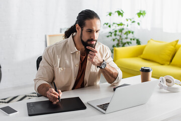 thoughtful hispanic retoucher touching beard while working with laptop and graphic tablet at home