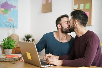 Happy gay couple kissing while using laptop in living room at home - LGBT love and technology...