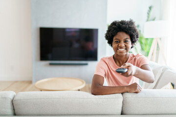 Cheerful cute woman sitting on sofa in bright living room changing tv channel