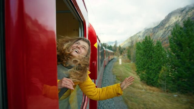 Young traveller lean from train. Woman hang out of train carriage window. Cinematic and inspiring travel blogger live motivational adventure. Happy young woman on train vacation, catch wind with hand