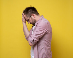 Bearded man with glasses is upset or has a headache, he closes eyes and holds forehead with hand. Yellow background.