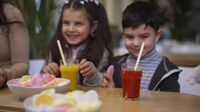 Waitress hands serving yellow and red juices for cheerful Middle Eastern boy and girl sitting at table in seafood restaurant. Positive laughing brother and sister enjoying family leisure. Slow motion