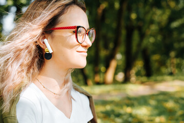 Profile portrait of young smiling woman listening music with portable wireless earphones outdoors, copy space. Hipster girl with glasses enjoying sound in headphones. Selective focus on audio device