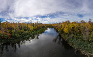 Fototapeta na wymiar Aerial view of brightly colored autumn trees surrounding a large calm river and a sky full of white clouds with blue patches. 
