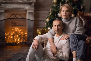 Fototapeta premium beautiful adult woman with a man sit on an armchair and hug near a christmas tree and fireplace