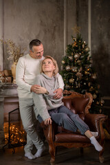 beautiful adult woman with a man sit on an armchair and hug near a christmas tree and fireplace