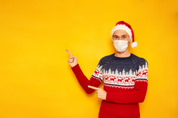 Fototapeta na wymiar man in Christmas sweater, medical mask, red Santa Claus hat points with his index finger at an empty area for New Year advertising. isolated yellow background with copy space concept - people, holiday