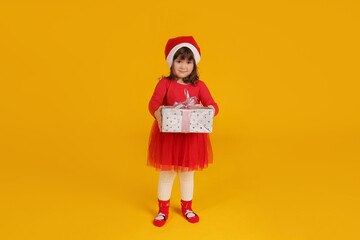 a little beautiful girl in a red Santa Claus hat dress holds a gift box for the new year in her hands