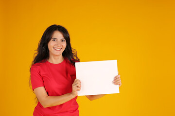 Fototapeta na wymiar beautiful happy brunette girl with long hair in a red T-shirt holding a white sheet of paper yellow background