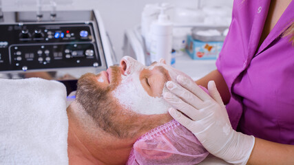 Obraz na płótnie Canvas Cosmetologist makes a man a facial therapeutic. Middle-aged man in a spa for beauty treatments. Beautician extrude from bottle with moisturizing cream on his hand to apply it to the male face.