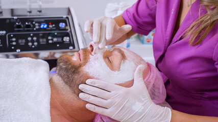  Cosmetologist makes a man a facial therapeutic. Middle-aged man in a spa for beauty treatments. Beautician extrude from  bottle with  moisturizing cream on his hand to apply it to the male face.