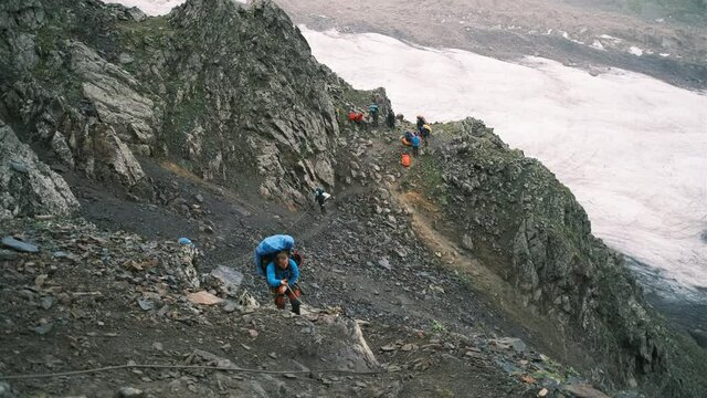 Climbers take turns walking one after another in the mountains holding on to the rope. A group of tourists with backpacks with equipment on the rocks are ferried along the railing
