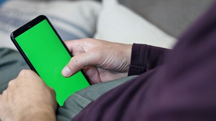 Close-up, man's hand touches and scroll with his finger while holding an Smartphone with green screen. Man lying on the sofa at home.