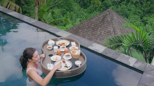 travel woman having breakfast in swimming pool enjoying exotic food at luxury hotel spa with view of tropical jungle at sunrise. A girl in a swimsuit eats food from a basket with a floating breakfast.