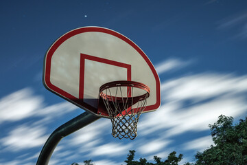 A long exposure photograph of a dirty orange and white playground basketball hoop, rim and net with motion blurred clouds in a dark blue night sky in the background. © Joseph Kirsch