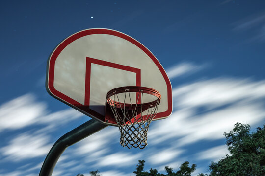 A long exposure photograph of a dirty orange and white playground basketball hoop, rim and net with motion blurred clouds in a dark blue night sky in the background.