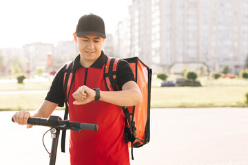 Fototapeta na wymiar Positive deliveryman worker employee deliver online order client customer. Smiling man courier food delivery with red thermal backpack walks street with electric scooter