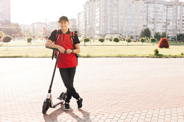 Portrait of deliverman standing with electric scooter with red thermal backpack looking at camera. E-Scooter rider eco transport. Modern urban alternative transport. Excellent delivery