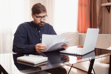 Fototapeta premium Entrepreneur in eyeglasses works with a laptop and keeps a document in a home office. Man holding paper documents, chatting online with clients on laptop at workplace.