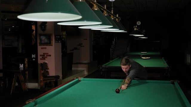 Close-up of a man playing pool in a dark bar, he takes aim and prepares to hit the ball with a cue. Playing Billiards on the green table.