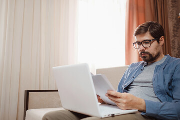 Entrepreneur in eyeglasses works with a laptop and keeps a document in a home office. Man holding paper documents, chatting online with clients on laptop at workplace.