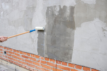 A worker applies the primer to the wall with a long-handled roller. A man's hand rolls a primer...