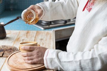 Ethnic linen shirt on the man. A man with a beard in the kitchen pours honey into a cup. Cooking dessert in a rustic kitchen.