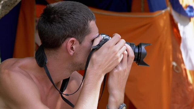 Young Photographer Resting near Tents Photographs Landscapes on a Riverbank. Unshaven male, a tourist, with a naked body, takes pictures of beach, forest, using a professional camera. Travel concept.