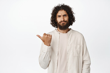 Portrait of arab guy pointing left, sulking and frowning, disapprove smth bad, dislike promo, standing over white background