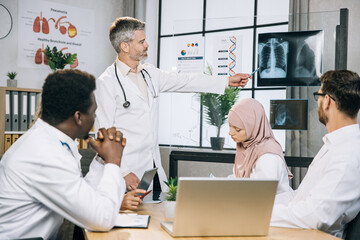 Obraz premium Qualified male doctor standing at boardroom and pointing on x ray scan while having seminar with multiracial practitioners. Team of therapists having meeting about progress in medical researches.