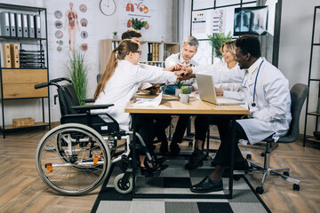 Fototapeta na wymiar Multiracial doctors with one woman using wheelchair holding their hands together in circle during successful meeting at office. Concept of medicine, science and cooperation.