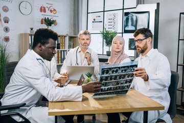 Five qualified mixed race doctors in uniform sitting at desk and looking at x ray scan. Group of therapists with one colleague who using wheelchair discussing ways of patient treatment.
