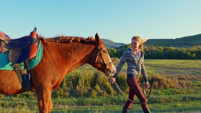 European long-haired girl leads chestnut horse by reins along hill. Panorama, landscape with mountain, wooded steppe. Young woman going with palfrey in countryside. Horsewoman walks bay stallion