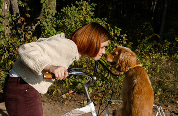 Happy young woman on the bicycle is cuddling, touching noses with her dog that sits in the basket.