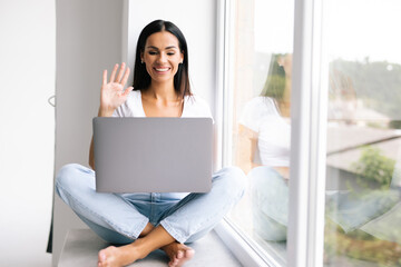 Working from home concept. Young woman using laptop for online video call or web conference with boss while sitting on windowsill, copy space