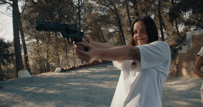 Young brunette woman with tattoo on hand aim at a target and shooting with air revolver in private forest of rustic house on summer evening at sunset