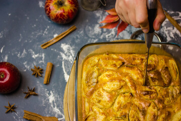 Woman holding a knife in hand, cutting a freshly baked apple pie in glass baking dish. Ingredients for baking - red apples, aniseed spices, cinnamon sticks, dusted flour top view. Thanksgiving dessert