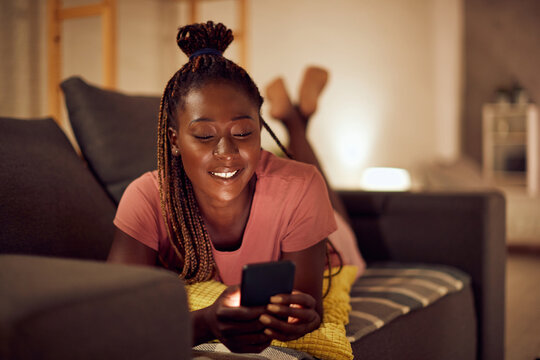 Happy African American woman types text message on cell phone while relaxing on sofa.