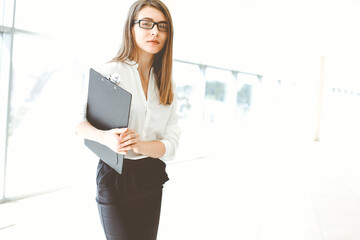 Beautiful female specialist with clipboard standing in modern office and smiling charmingly. Working on design, data analysis, plan strategy. Business people concept