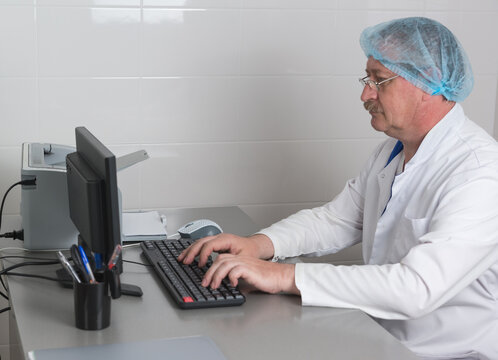 An elderly doctor in glasses and medical clothes is sitting at desk in the office and typing the conclusion of the examination on computer